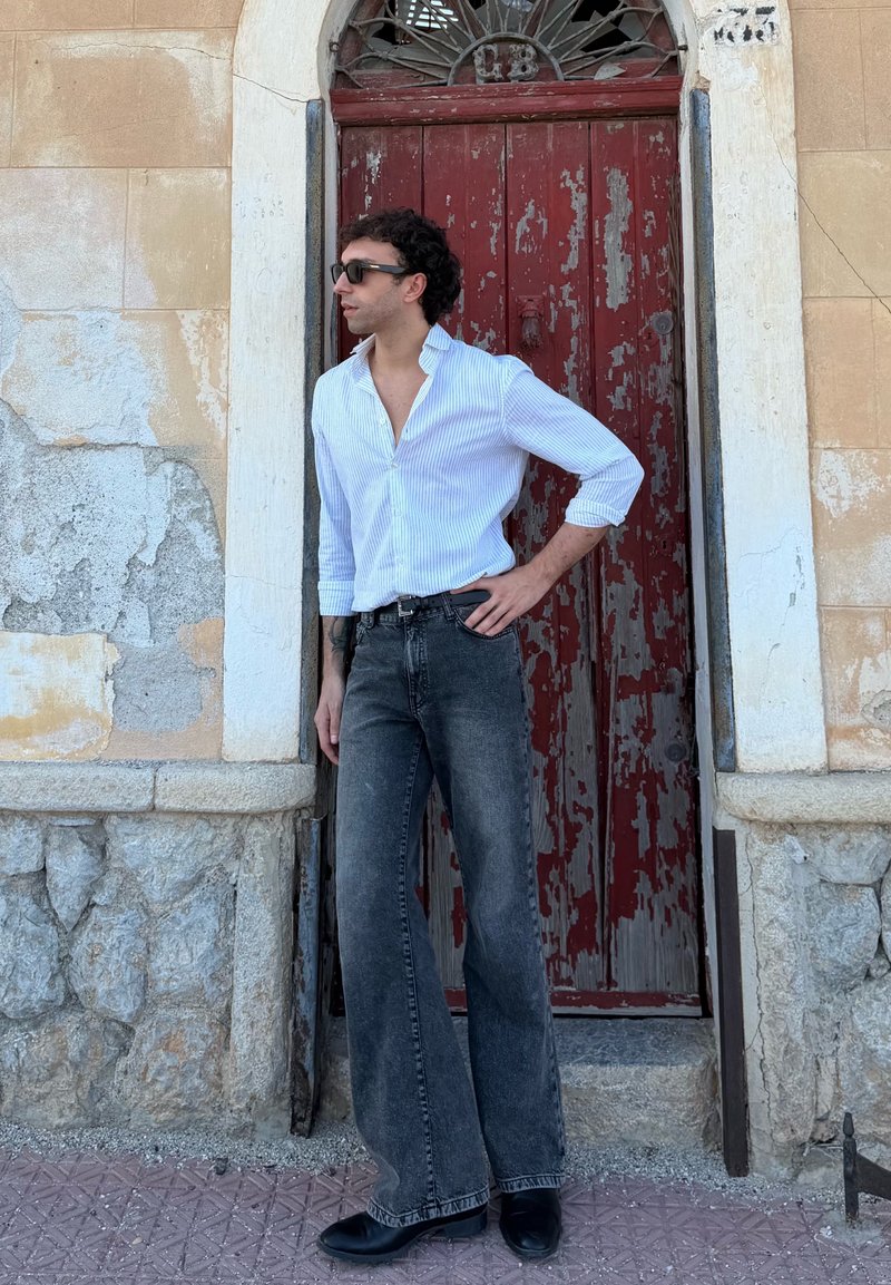 White and blue striped shirt, black high-waisted wide-leg jeans, black shoes; standing next to a weathered red door and stone wall.