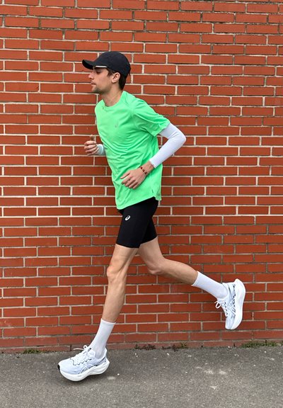 Joven corriendo con camiseta verde brillante, pantalones cortos negros, zapatillas blancas y gorra negra frente a una pared de ladrillos rojos.