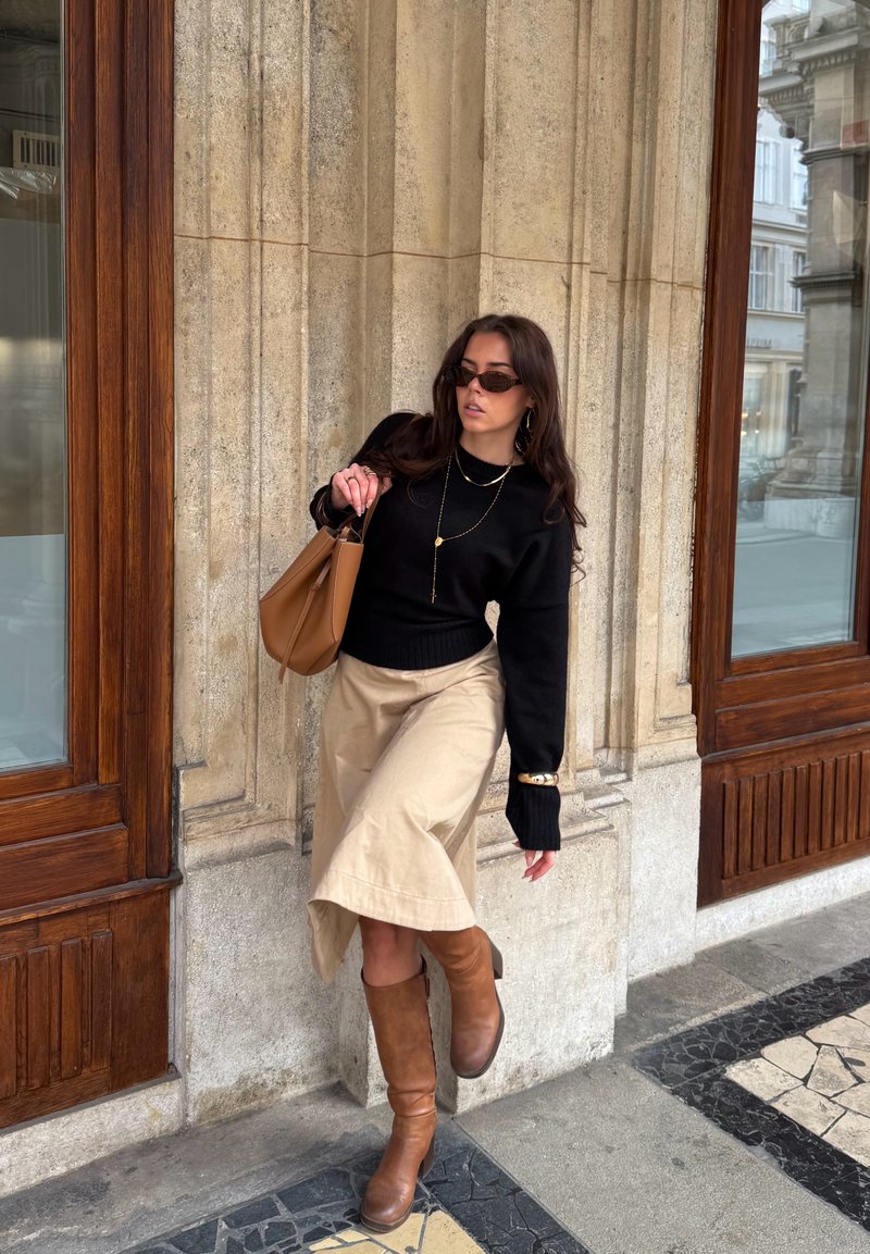 Fekete pulóvert, bézs szoknyát és barna csizmát viselő nő barna táskával, kőfal mellett állva egy fa ajtókeretek közelében.