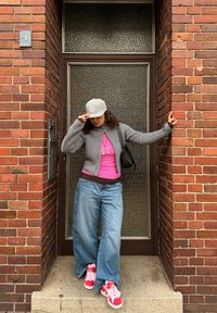 Cardigan en tricot gris, haut rose, jean large en denim et baskets rouges avec lacets roses. Debout devant un mur en verre texturé et en briques.