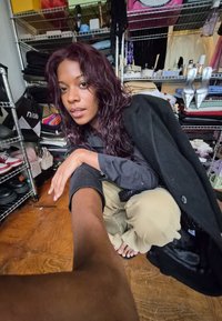 Fekete kabát gombos ing felett, bézs nadrág és mezítláb. A háttérben cipők, táskák és összehajtogatott ruhák találhatók a polcokon.