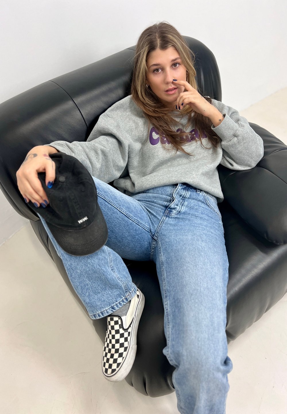 Grey sweatshirt with purple text, relaxed blue jeans, black and white checkered slip-on shoes, sitting on a black leather sofa, holding a cap.