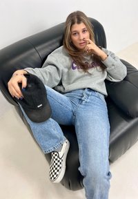 Grey sweatshirt with purple text, relaxed blue jeans, black and white checkered slip-on shoes, sitting on a black leather sofa, holding a cap.