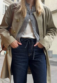 Trench coat beige sopra un cardigan grigio, una maglietta bianca a costine e jeans in denim blu scuro. Tasche in evidenza, borsa a tracolla marrone visibile.