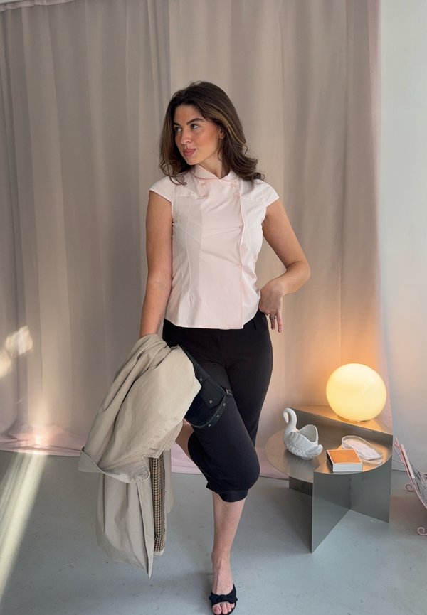 Una mujer posa con una blusa rosa pálido y pantalones negros, sosteniendo un abrigo beige junto a una mesa con una lámpara encendida.