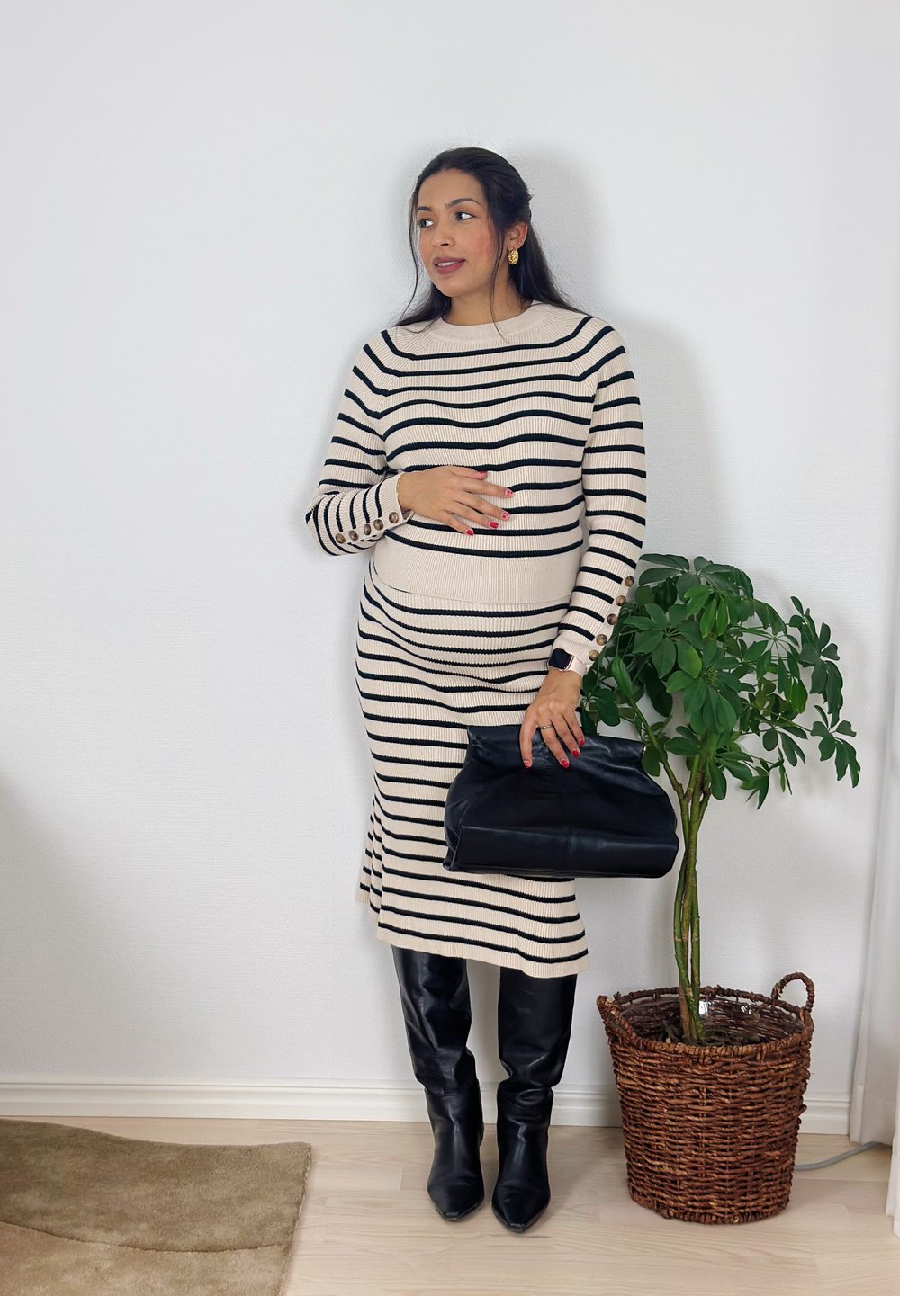 Femme en robe beige à rayures noires, bottes noires jusqu'aux genoux, tenant un sac à main noir, debout à côté d'une plante verte en pot contre un mur blanc.