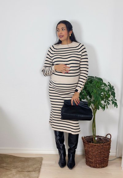 Mujer con vestido beige y negro a rayas, botas negras hasta la rodilla, sosteniendo un bolso negro, de pie junto a una planta verde en maceta contra una pared blanca.