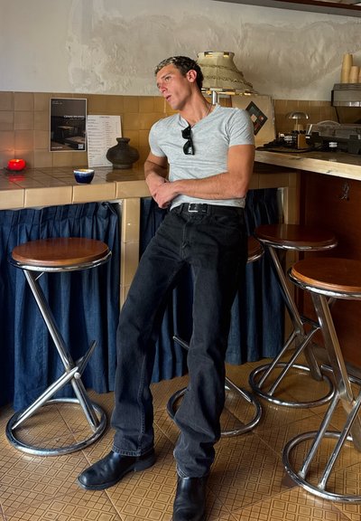 Hombre con camiseta gris y vaqueros negros apoyado en un mostrador de bar con azulejos con taburetes, tazas y lámpara en un ambiente acogedor de cafetería.