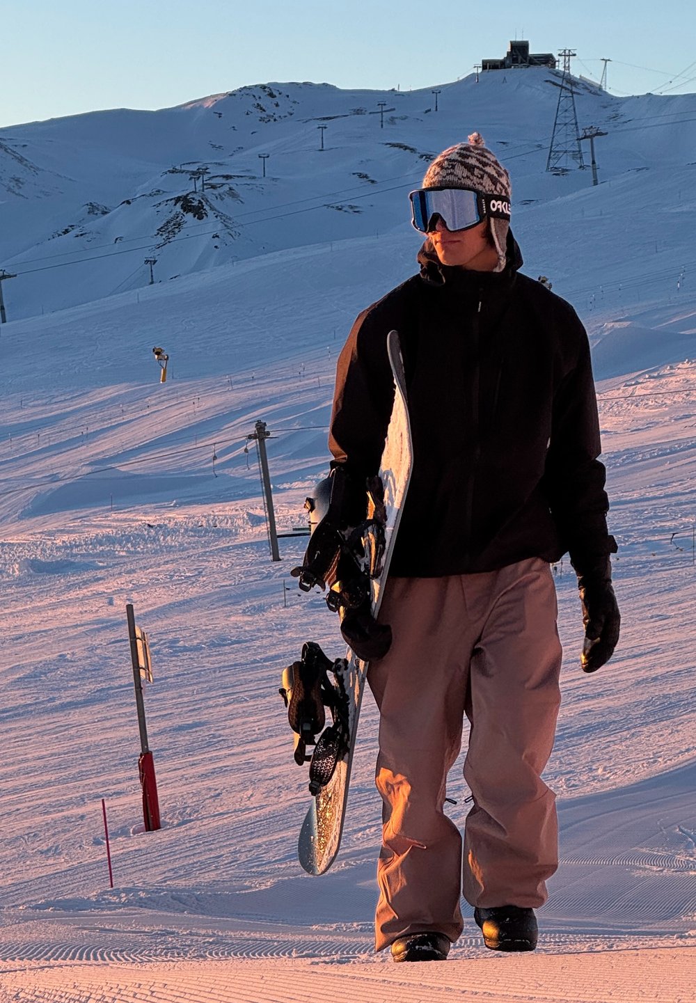 Deskar snowboarder drži snowboard, oblečen v črno jakno in svetlo rjave snežne hlače, s pisano kapo in odsevno očali, na zasneženi površini.