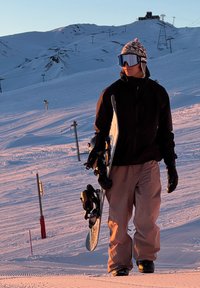 Deskar snowboarder drži snowboard, oblečen v črno jakno in svetlo rjave snežne hlače, s pisano kapo in odsevno očali, na zasneženi površini.