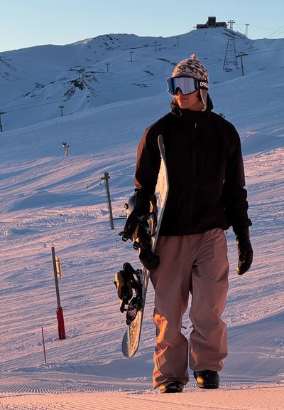 Snowboarder sosteniendo una tabla de snowboard, vistiendo una chaqueta negra y pantalones de nieve marrón claro, con un gorro de patrones y gafas de sol reflectantes, en un terreno nevado.
