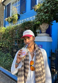 White shirt, brown and cream checked scarf, red and white cap, holding a drink with a blue straw, blue wall with plants in the background.
