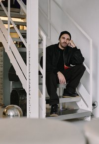Hombre sentado en escaleras blancas, vistiendo una chaqueta negra y un gorro. Pantalones negros y zapatos con una suela texturizada. Ambiente interior minimalista.