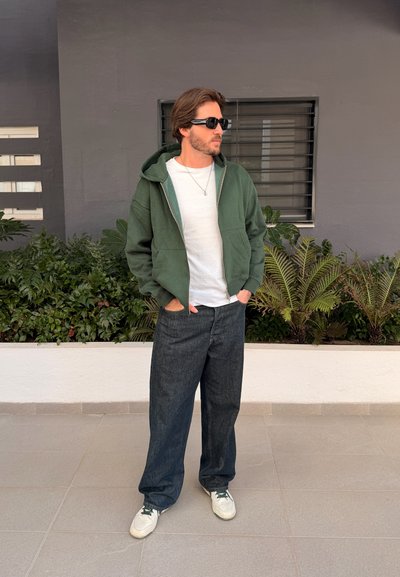 Hombre con gafas de sol, vestido con sudadera verde con cremallera, camiseta blanca, pantalones negros holgados y zapatillas blancas, de pie cerca de plantas y una pared gris.