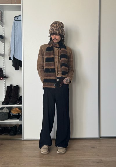 Persona con un sombrero de estampado de leopardo, cárdigan marrón, bufanda a rayas, pantalones anchos negros y zapatos beige, de pie junto a una pared blanca cerca de estanterías con ropa.