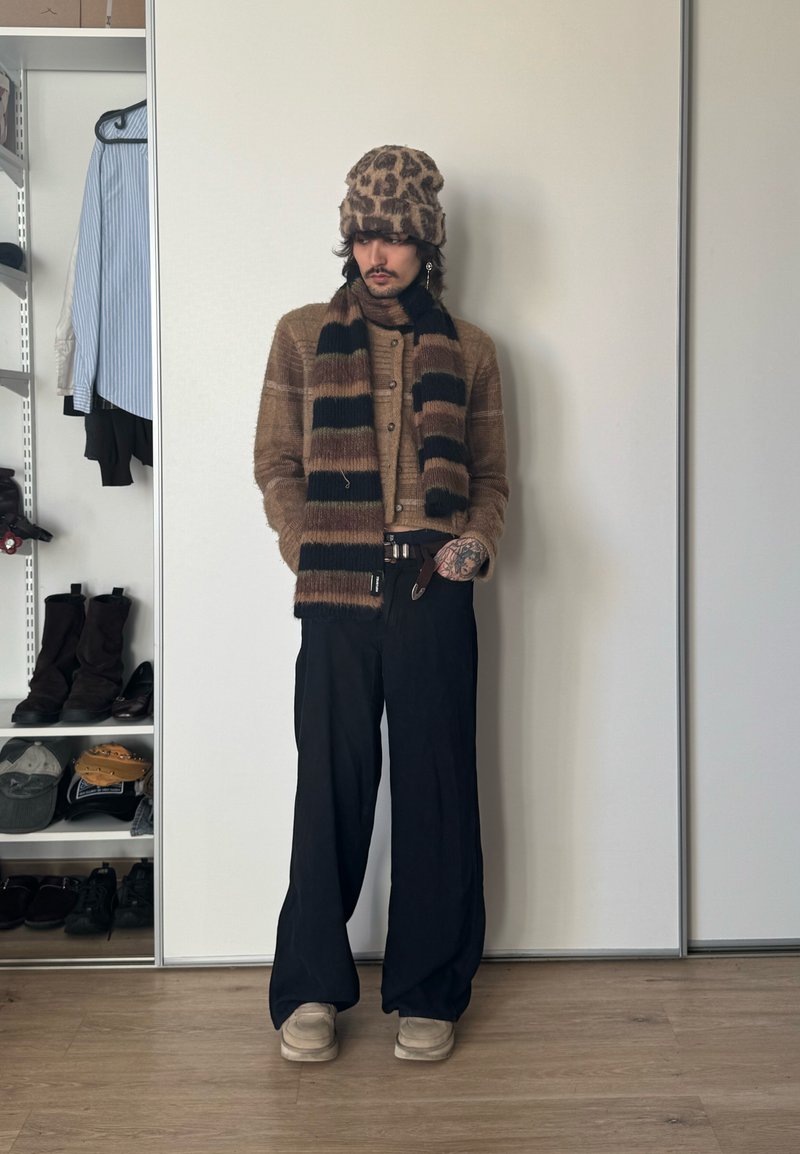 Inimene, kes kannab leopardimustrilist mütsi, pruuni kardigani, triibulist salli, laiu musti pükse ja beeže jalatseid, seisab valge seina ääres, lähedal riietega riiulitele.