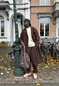 Casaco trench castanho sobre um suéter creme com o texto "QUO", calças castanhas, sapatos rosa e uma mala cinza. Ambiente de rua com bicicletas.