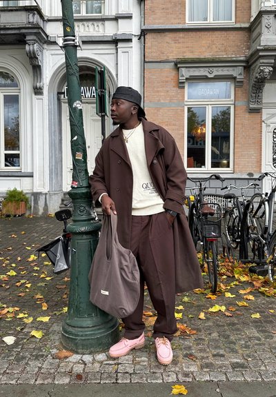 Abrigo trenca marrón sobre un suéter crema con el texto "QUO", pantalones marrones, zapatos rosas y un bolso gris. Entorno urbano con bicicletas.