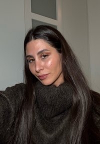 Ciemnoszary, oversize'owy sweter z wysokim kołnierzem, gładką fakturą, noszony z długimi ciemnymi włosami, naturalnym makijażem i neutralnym kolorem ust.