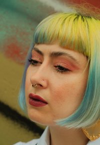 Kort blå og gul hår, detaljeret øjenmakeup med shimmer, septum ring, guld næsepiercing og mørkerøde læber mod en tekstureret baggrund.