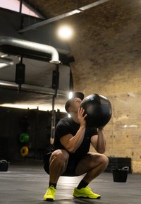 Palla medica nera tenuta all'altezza del petto da una persona in posizione accovacciata. Scarpe da ginnastica giallo brillante, maglietta nera, ambiente palestra minimalista con pareti in mattoni.
