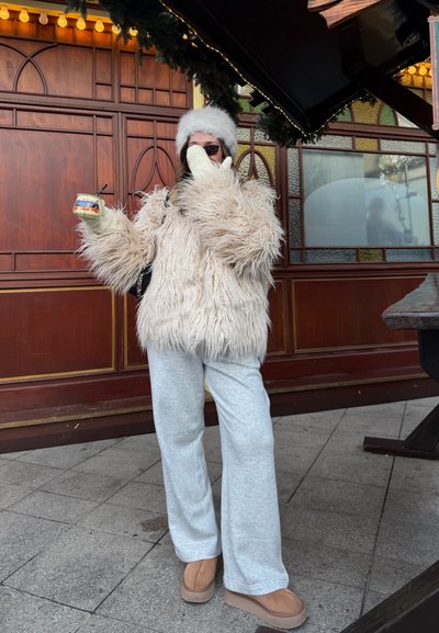 Chaqueta de piel sintética beige con mangas largas, pantalones deportivos grises y botas color canela. Sosteniendo una taza pequeña. Fondo con paneles de madera y luces festivas.