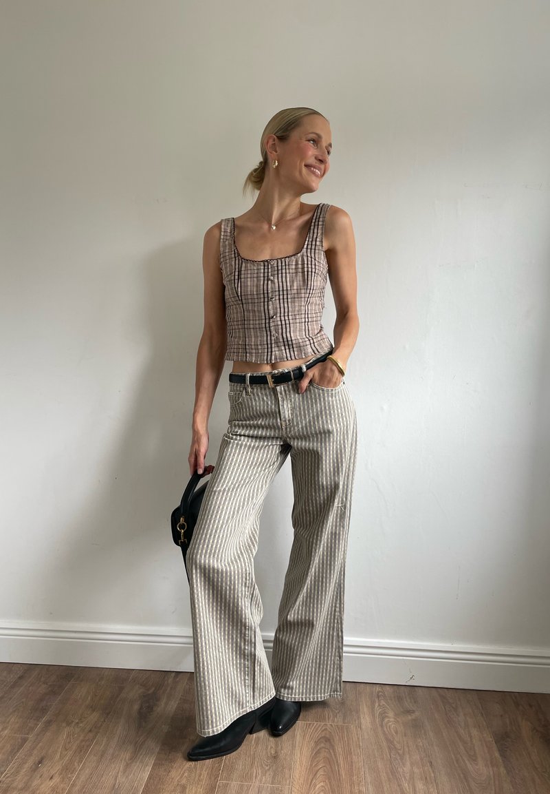 Femme debout à l'intérieur portant un haut sans manches à carreaux, un pantalon large rayé, des bottes noires et tenant un sac à main noir.