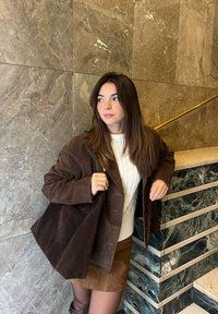 Jeune femme en veste marron et jupe avec un pull blanc se tient près d'un mur en marbre et d'une rampe d'escalier, regardant vers la gauche.