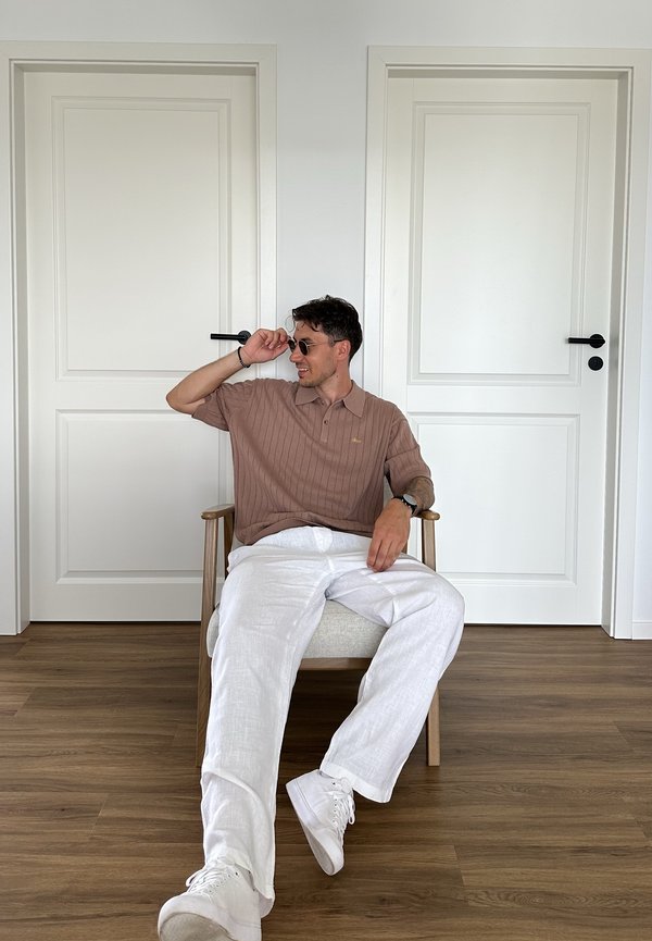 Uomo con occhiali da sole, camicia marrone e pantaloni bianchi seduto su una sedia di legno con un cuscino chiaro tra due porte bianche su un pavimento in legno.