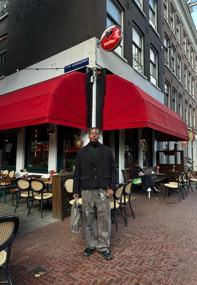 Una persona está de pie frente a un café, vistiendo un blazer negro, pantalones grises texturizados y sosteniendo una bolsa gris. Se pueden ver un toldo rojo y mesas de café.