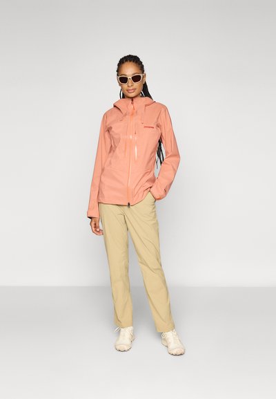 Chaqueta impermeable ligera color durazno con un diseño ajustado, cierre de cremallera y capucha ajustable; combinada con pantalones beige y zapatillas neutras.