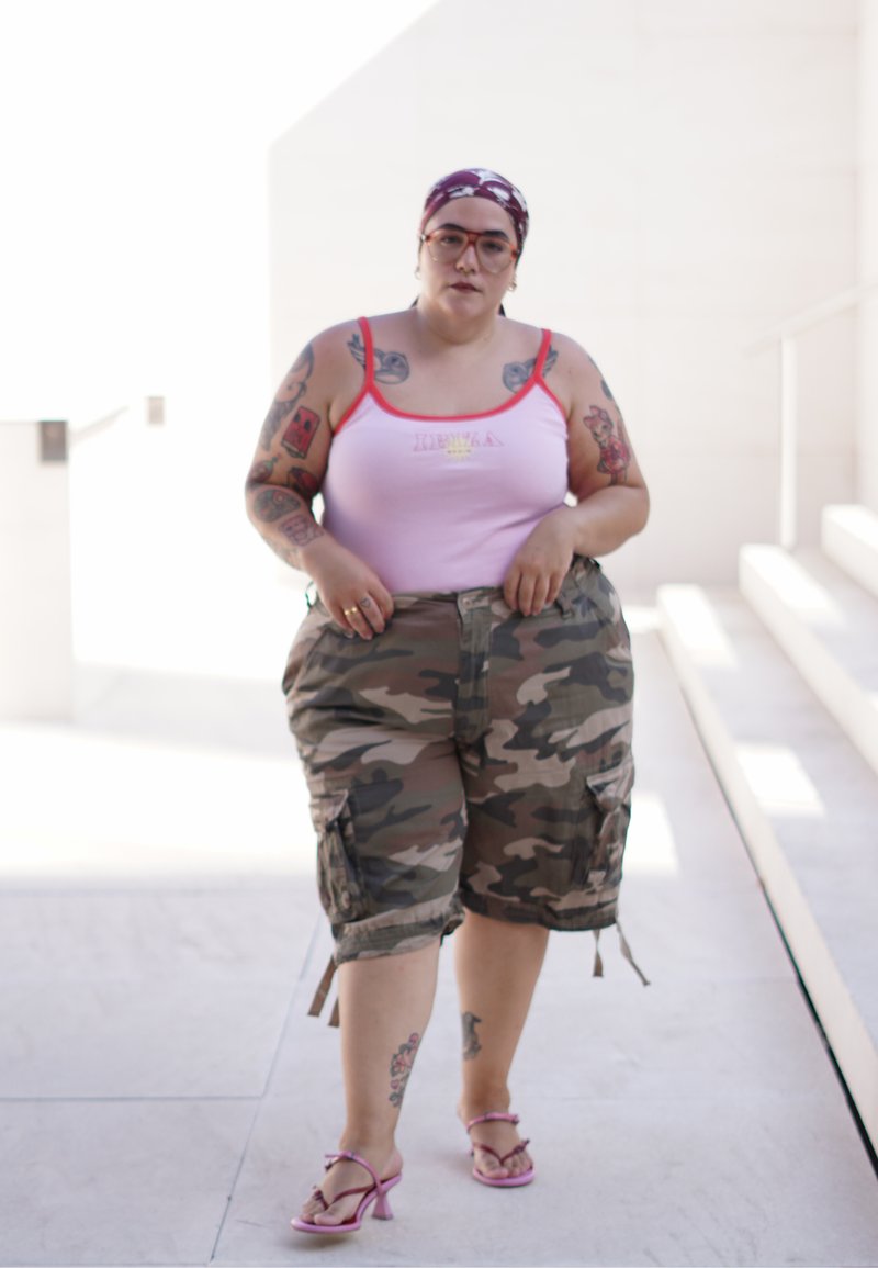 Personne portant un débardeur rose, un short camouflage, des talons roses et un foulard, marchant dans un environnement urbain extérieur lumineux.