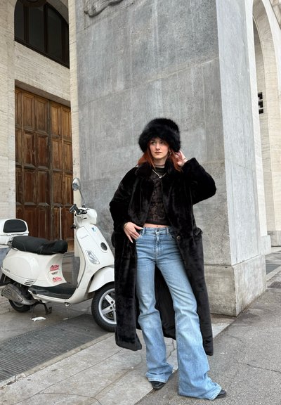 Abrigo de piel sintética negro con botones, combinado con una blusa negra transparente y vaqueros acampanados azul claro. Un gorro de piel negro completa el conjunto. Scooter blanco cerca.
