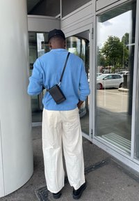 Lys blå ribbet genser, hvite vide bukser, svart crossover-veske og svarte platåsko, står nær en glassinngang.