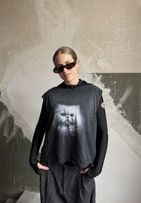 Čierne skrátené tričko s potlačenou bielou mačacou tvárou a textom pod ňou. Nosené cez čierne tričko s dlhým rukávom; v kombinácii s textúrovanými sivými nohavicami.