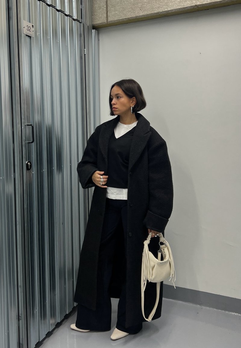 Long manteau en laine noire porté sur un haut noir et une chemise blanche, associé à un pantalon noir à jambes larges et un sac à main en cuir crème ; boucles d'oreilles anneaux argentés.