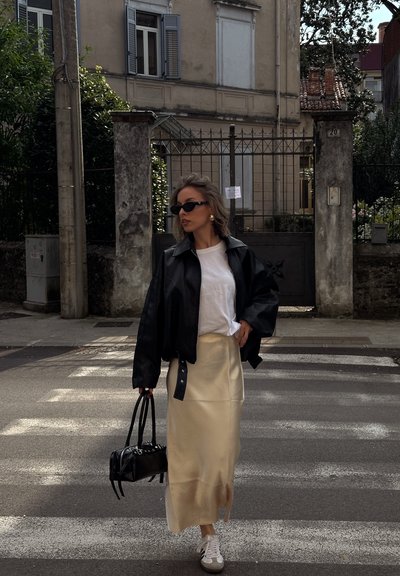 Mujer con chaqueta de cuero negra, camiseta blanca, falda beige y zapatillas deportivas caminando por un paso de peatones, sosteniendo un bolso negro, frente a un edificio cerrado con rejas.