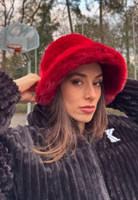 Femme ajustant un chapeau rouge vif et duveteux, portant une veste noire texturée, avec un panier de basket-ball en plein air et des arbres en arrière-plan.