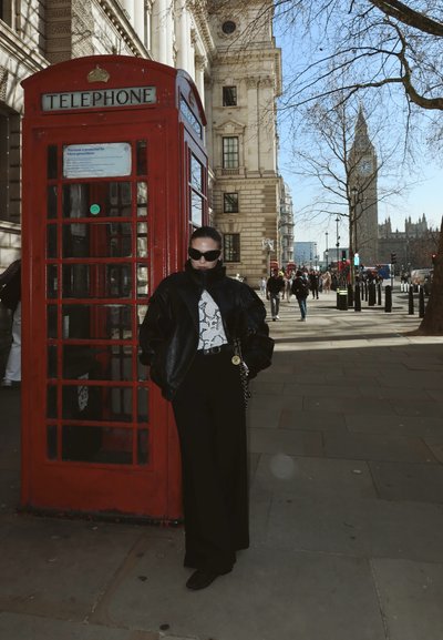 Persona con chaqueta negra y gafas de sol de pie junto a una cabina telefónica británica roja, con el Big Ben visible al fondo.