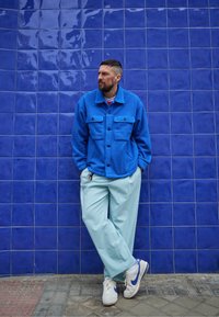 Casaco de fleece azul com dois bolsos frontais, calças largas azul claro, e sapatilhas brancas com um detalhe azul, contra uma parede azulejada azul.