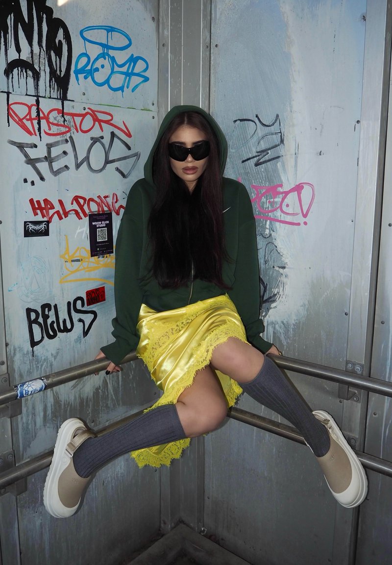 Frau in dunklem Hoodie, Sonnenbrille, gelbem Rock und knielangen Socken, die mit ausgestreckten Beinen auf Metallstangen in einer mit Graffiti bedeckten industriellen Ecke sitzt.