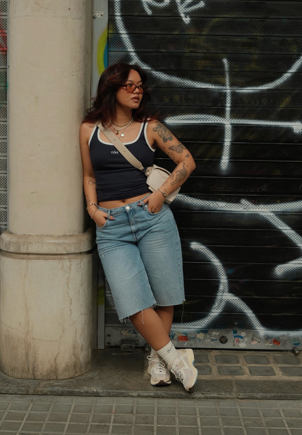Camisola de alças de riscas azul-marinho com borda branca, calções de ganga azul claro, mala de ombro creme e sapatilhas multicoloridas. Tatuagens nos braços visíveis.