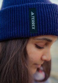 Fialová pletená čepice s žebrovanou texturou a černou štítkem s logem "TERREX" umístěným na boku. Nese ji osoba směřující hlavou dolů.