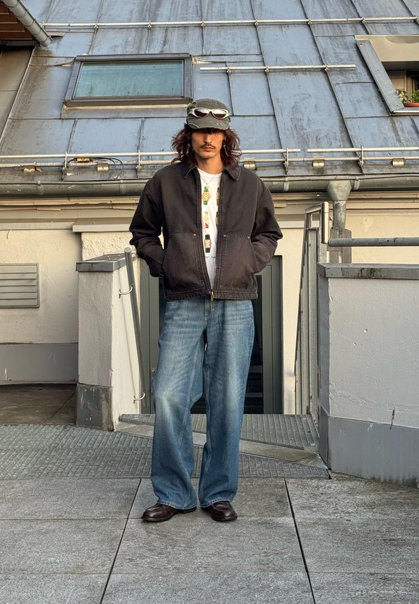 Homme aux cheveux longs et à la moustache, debout dans les escaliers du toit, portant une veste foncée, une chemise blanche, un jean bleu ample, une casquette avec des lunettes de soleil posées dessus.