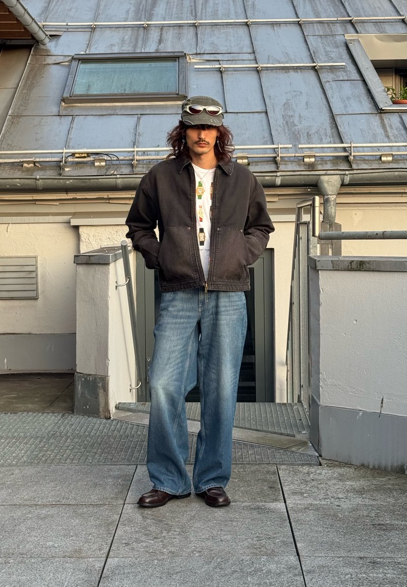 Man with long hair and mustache stands on rooftop stairs wearing dark jacket, white shirt, loose blue jeans, cap with sunglasses resting on it.