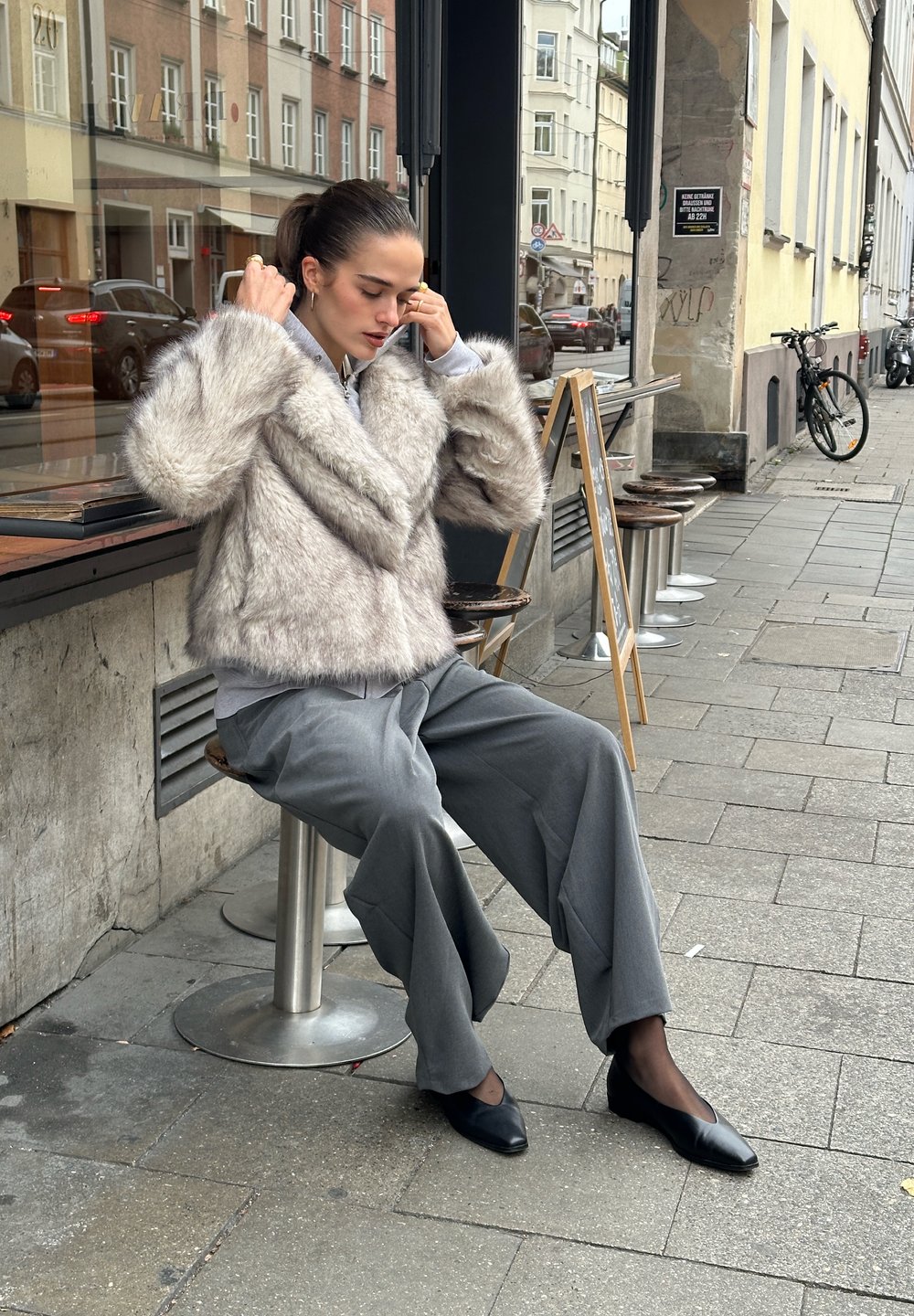 Faux fur jakke i lysegrått, kort design. Kombinert med løse grå bukser og svarte leiligheter. Sitter på en krakk utenfor en kafé.