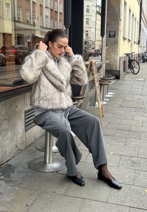 Chaqueta de pelo sintético en gris claro, de diseño corto. Combinada con pantalones grises holgados y bailarinas negras. Sentada en un taburete fuera de un café.