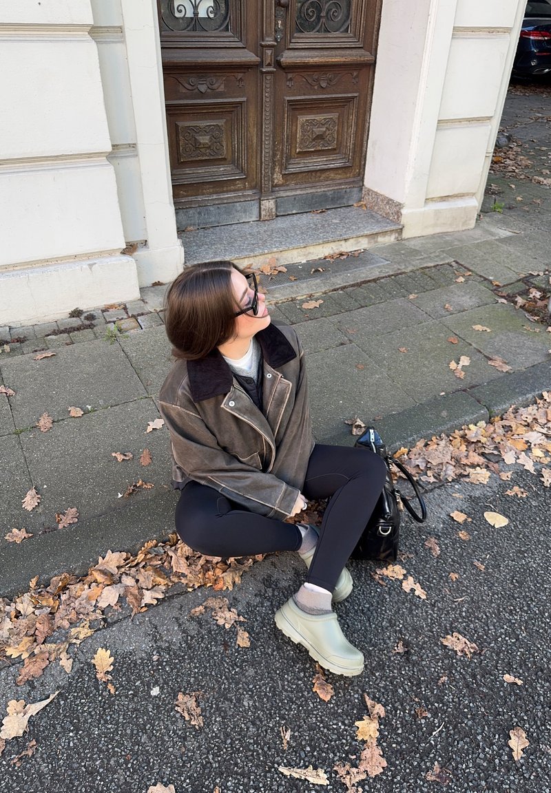 Leggings pretas ajustadas, casaco de couro castanho oversized com gola escura, tamancos verde claro e uma bolsa preta, sentada num caminho pavimentado.