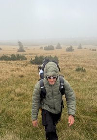 Muškarac u zelenoj jakni s kapuljačom i sunčanim naočalama hoda kroz maglovito polje s ruksakom na travnatome terenu s raspršenim grmovima.