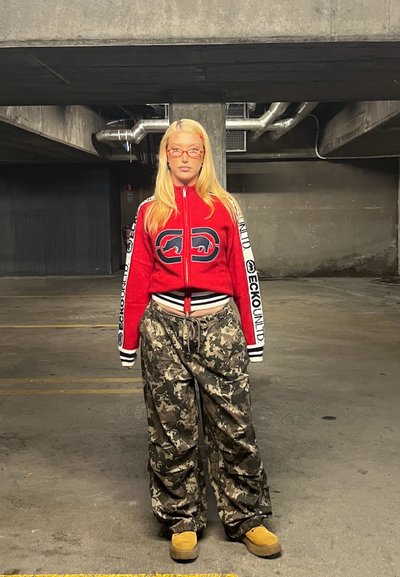 Chándal rojo con cremallera y acentos en blanco y negro, pantalones camuflados holgados y calzado beige. Lentes redondos notables y cabello largo y rubio.